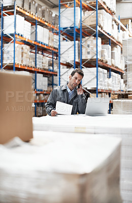 Buy stock photo A young man sitting in a warehouse taking orders on his cellphone and recording them on his laptop
