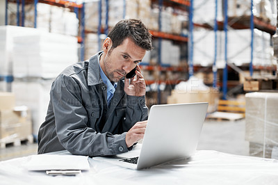 Buy stock photo A young man sitting in a storehouse taking orders on his cellphone and recording them on his laptop