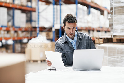 Buy stock photo A young man using his laptop and looking over his notes while working in a storehouse