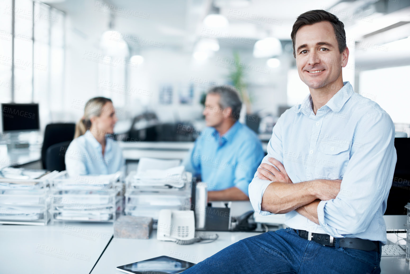 Buy stock photo Confident businessman sitting on a desk with his coworkers in the background