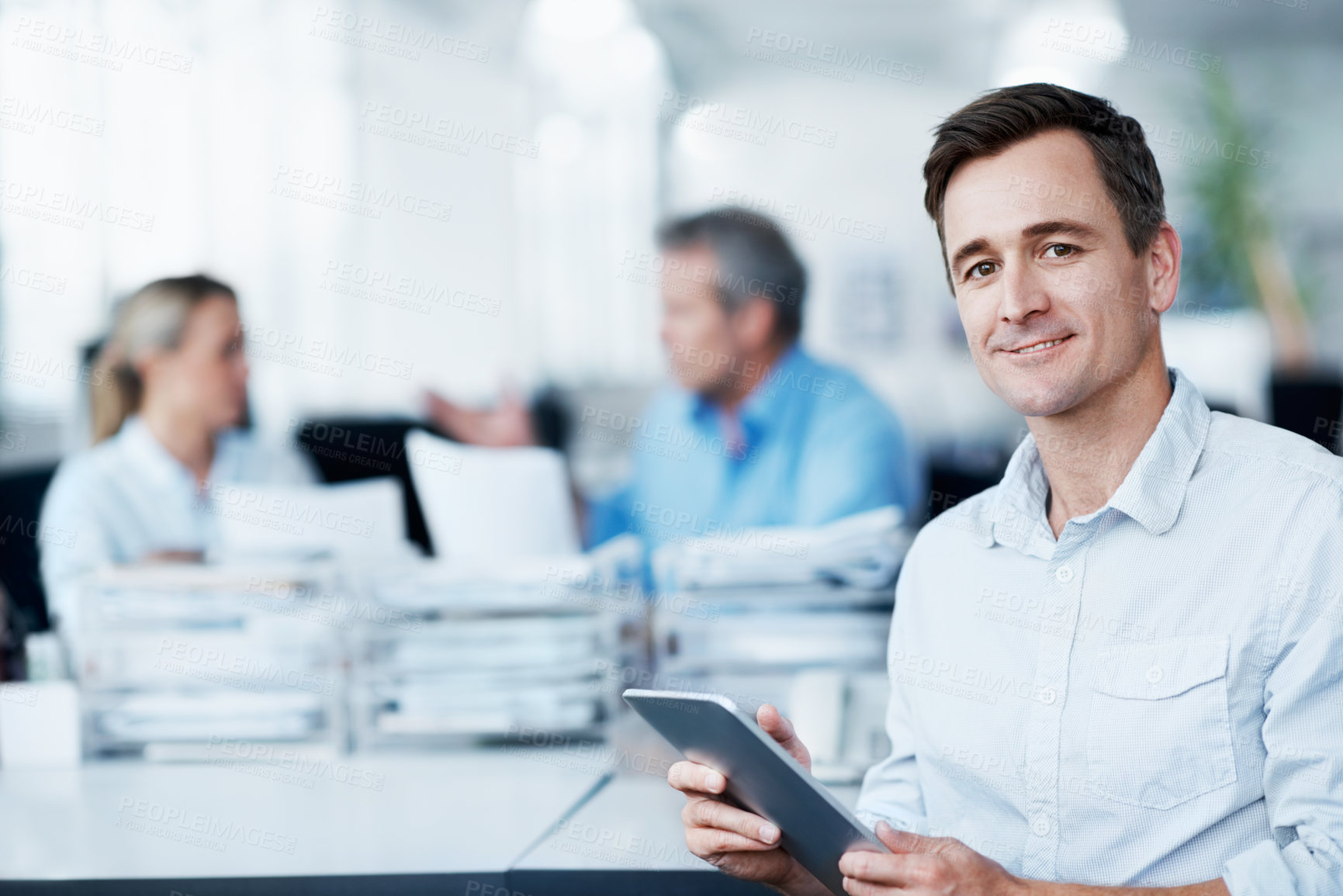 Buy stock photo Portrait of a handsome young man using his digital tablet while two colleagues converse in the background