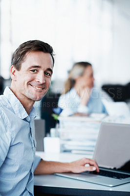 Buy stock photo Portrait of a young businessman working on his laptop with his colleague in the background