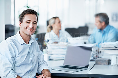 Buy stock photo Portrait of a young businessman sitting at his desk while his two coworkers talk in the background