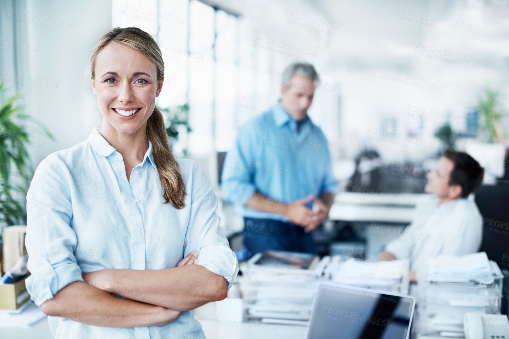 Buy stock photo Portrait of an attractive young businesswoman standing with her arms folded while her colleagues talk in the background