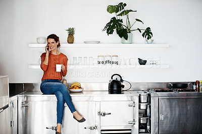 Buy stock photo Shot of a woman sitting on her kitchen counter drinking a coffee and talking on the phone