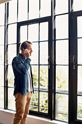 Buy stock photo Shot of a mature man talking on a cellphone while standing by windows in his apartment