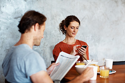Buy stock photo Shot of a couple eating breakfast at their kitchen table while reading the paper and using a cellphone