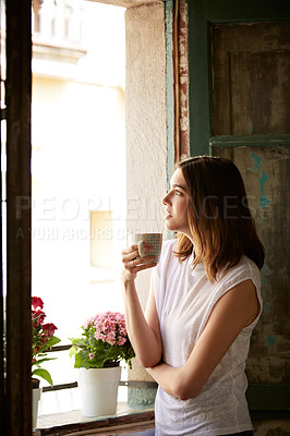 Buy stock photo Shot of a young woman drinking a cup of coffee while looking out of a window