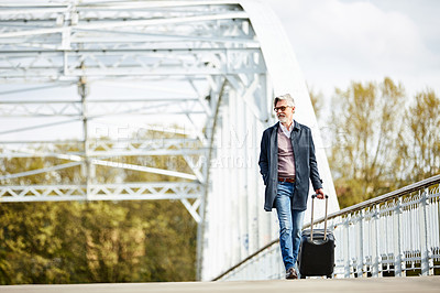 Buy stock photo Shot of a mature man pulling a suitcase while walking over a bridge in the city