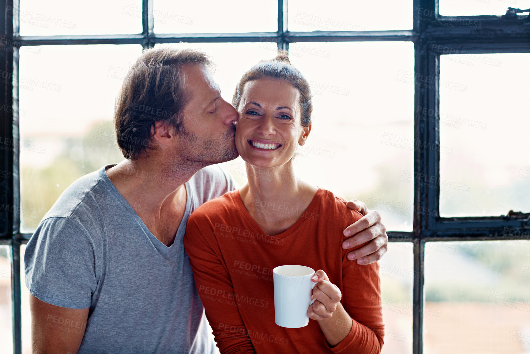 Buy stock photo Happy, couple and coffee with kiss at window for bonding together, connection and weekend break. Portrait, woman and mature man with affection in home for healthy marriage, love embrace and beverage
