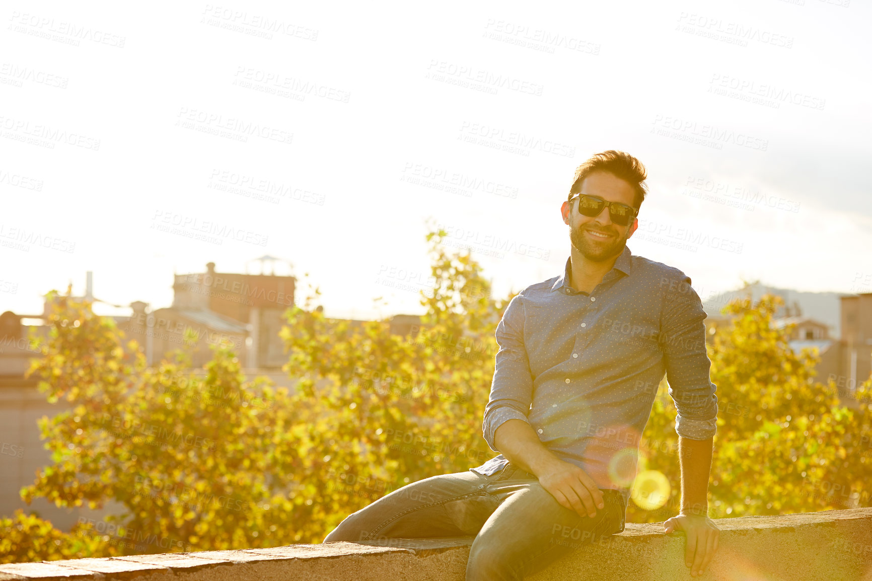 Buy stock photo Portrait of a smiling young man sitting on a rooftop at sunset with the city in the background