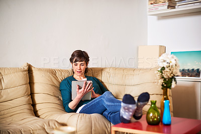 Buy stock photo Shot of a young woman sitting on her living room sofa using a digital tablet