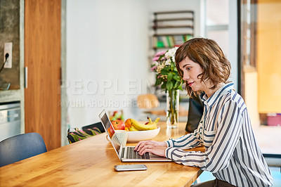 Buy stock photo Shot of a young woman sitting at her kitchen table working on a laptop
