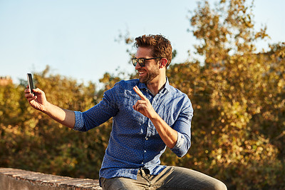 Buy stock photo Shot of a young man sitting on a wall taking a selfie with the city in the background