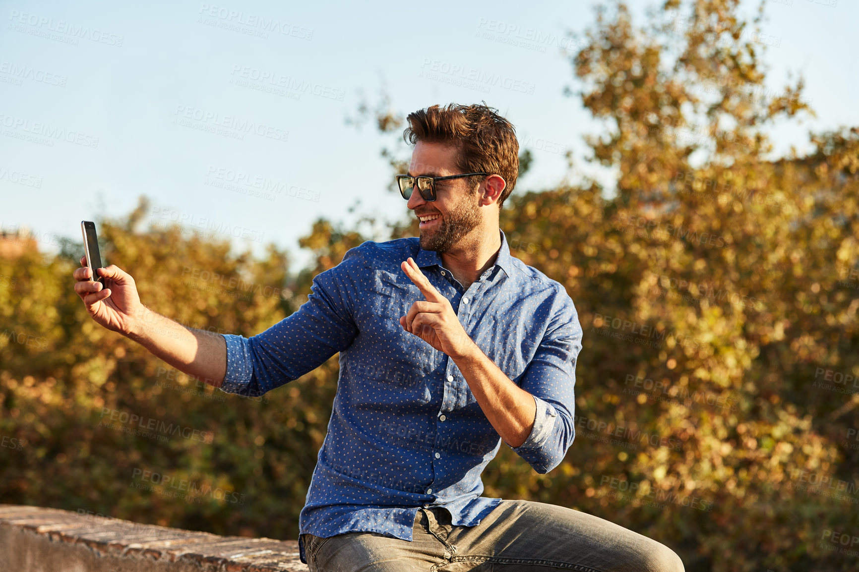 Buy stock photo Shot of a young man sitting on a wall taking a selfie with the city in the background