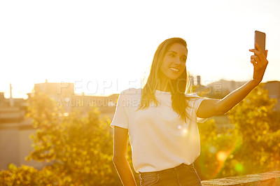 Buy stock photo Shot of an attractive young woman leaning on a wall taking a selfie with the city in the background