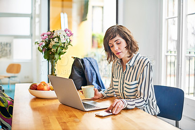 Buy stock photo Portrait of a young woman checking her cellphone while sitting at her kitchen table working on a laptop