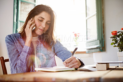 Buy stock photo Shot of an attractive young woman sitting by a window at home talking on a cellphone and writing on a notepad