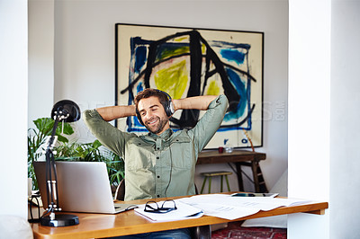 Buy stock photo Shot of a young man sitting at home with his hands behind his head wearing headphones and working on a laptop