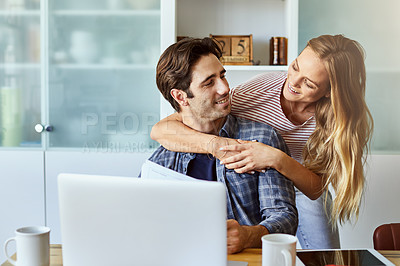 Buy stock photo Shot of a young woman hugging her husband while he works on his laptop at home