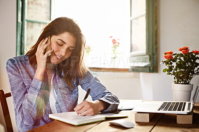 Buy stock photo Shot of an attractive young woman sitting by a window at home talking on a cellphone and writing on a notepad