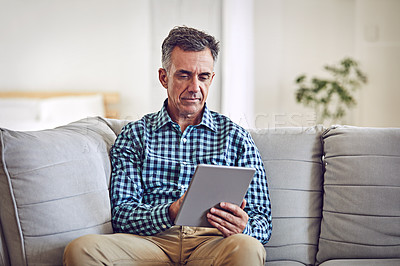 Buy stock photo Shot of a handsome and mature man using a tablet while sitting on the sofa at home