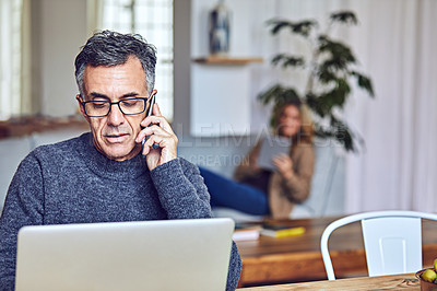 Buy stock photo Shot of a handsome and mature man working from home with his wife in the background