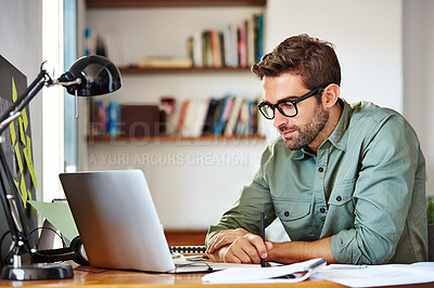 Buy stock photo Shot of a young man sitting at a table at home working on a laptop