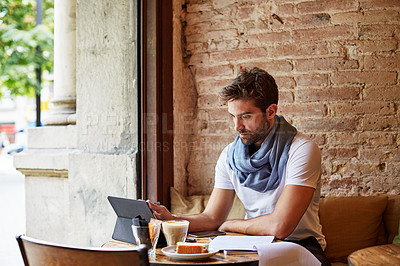 Buy stock photo Shot of a handsome young man using his digital tablet while sitting in a cafe
