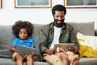 Buy stock photo Shot of a cheerful young father and son each browsing on a digital tablet while being seated on the couch at home