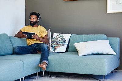 Buy stock photo Shot of a focused young man listening to music through headphones while being seated on a couch at home