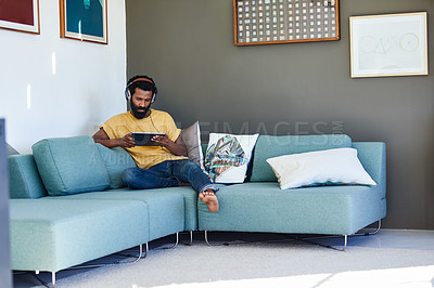 Buy stock photo Shot of a focused young man listening to music through headphones while being seated on a couch at home