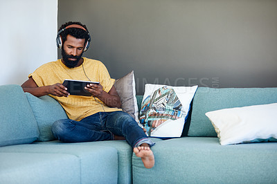 Buy stock photo Shot of a focused young man listening to music through headphones while being seated on a couch at home