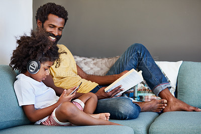 Buy stock photo Shot of a cheerful father and son listening to music and reading a book while relaxing on a couch at home