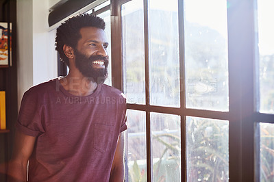 Buy stock photo Shot of a cheerful young man standing next to a window and looking through it while smiling brightly at home during the day
