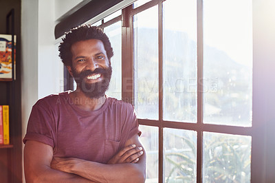 Buy stock photo Portrait of a cheerful young man standing next to a window with his arms folded inside at home during the day