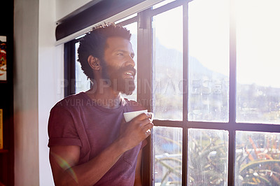 Buy stock photo Shot of a cheerful young man standing next to a window and looking through it while drinking a cup of coffee inside at home during the day