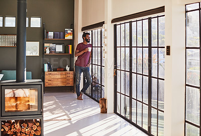Buy stock photo Shot of a cheerful young man standing next to a window and looking through it while drinking a cup of coffee inside at home during the day