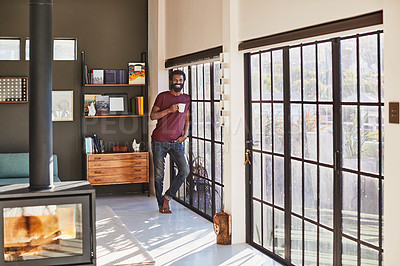 Buy stock photo Portrait of a cheerful young man standing next to a window and looking through it while drinking a cup of coffee inside at home during the day