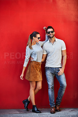 Buy stock photo Shot of a fashionable young couple leaning against a red wall outside
