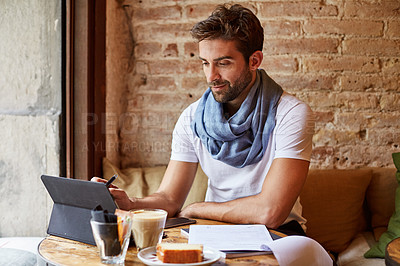 Buy stock photo Shot of a handsome young man using his digital tablet while sitting in a cafe