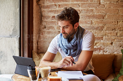 Buy stock photo Shot of a handsome young man using his digital tablet while sitting in a cafe
