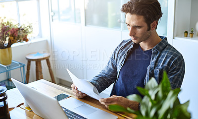 Buy stock photo Shot of a focused young man doing paperwork while being seated in front of his laptop at home