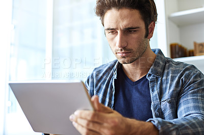 Buy stock photo Shot of a focused young man working on his digital tablet while being seated at home