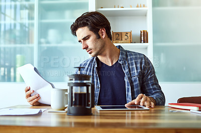 Buy stock photo Shot of a focused young man working on his digital tablet while being seated next to a table at home
