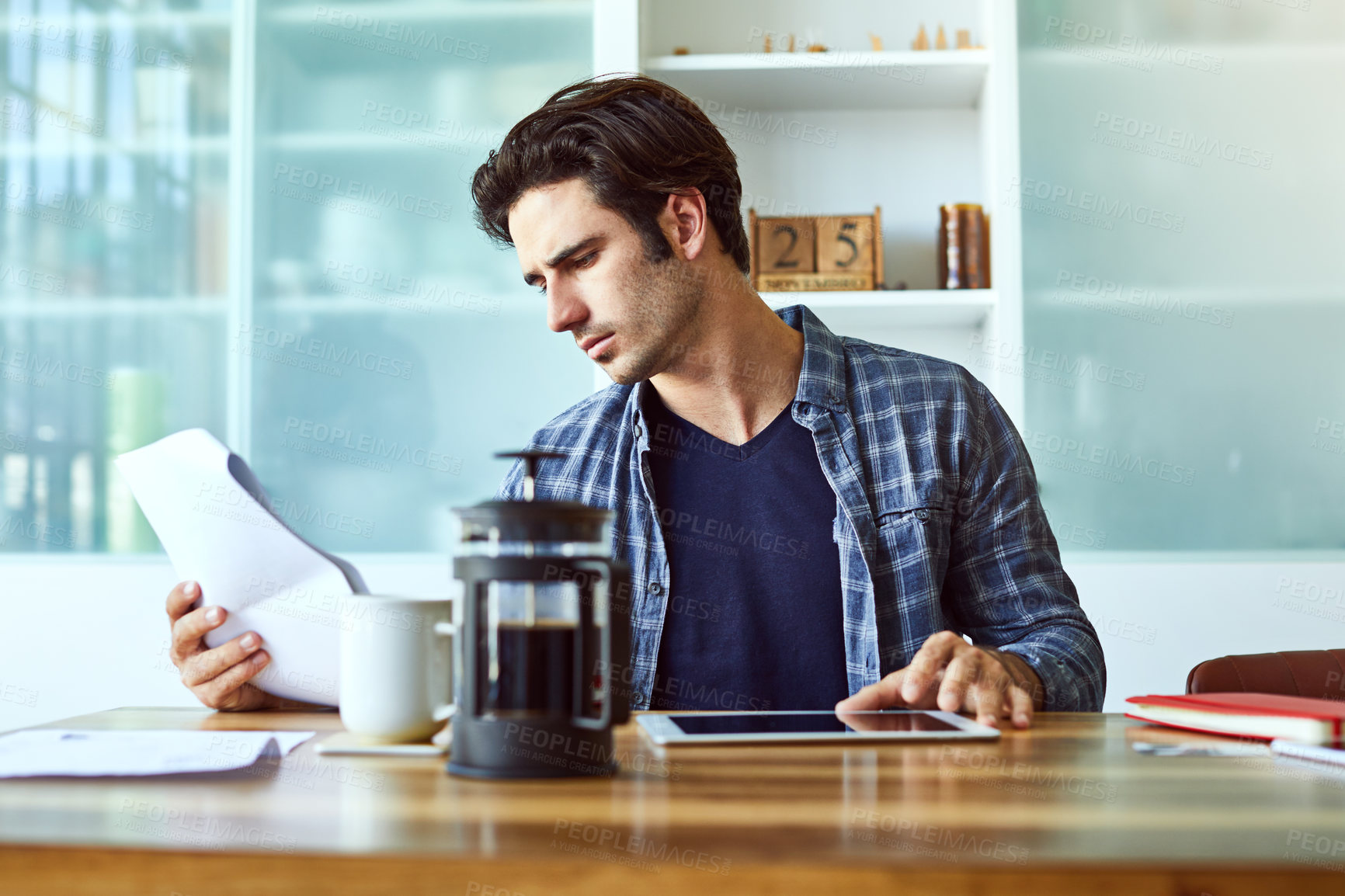 Buy stock photo Shot of a focused young man working on his digital tablet while being seated next to a table at home