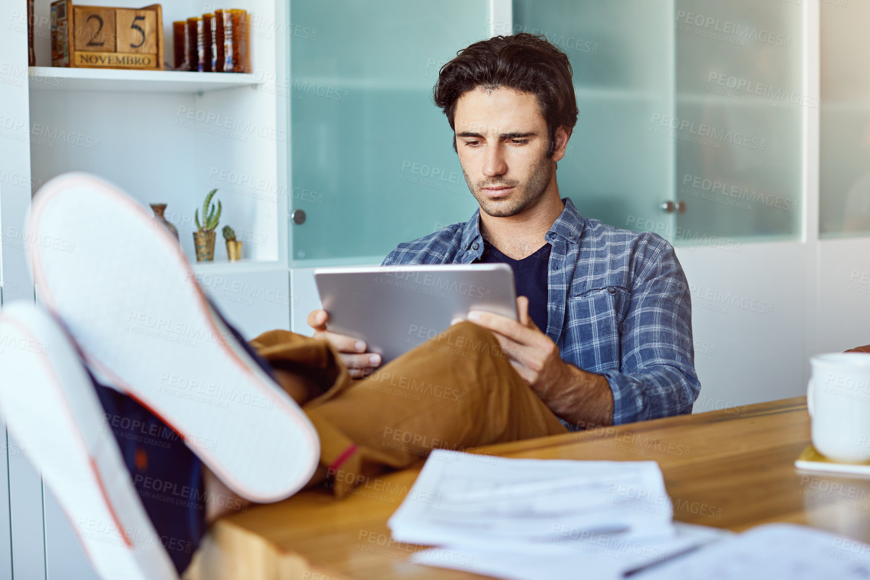 Buy stock photo Shot of a focused young man working on his digital tablet while being seated next to a table with his feet up at home