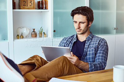 Buy stock photo Shot of a focused young man working on his digital tablet while being seated next to a table with his feet up at home