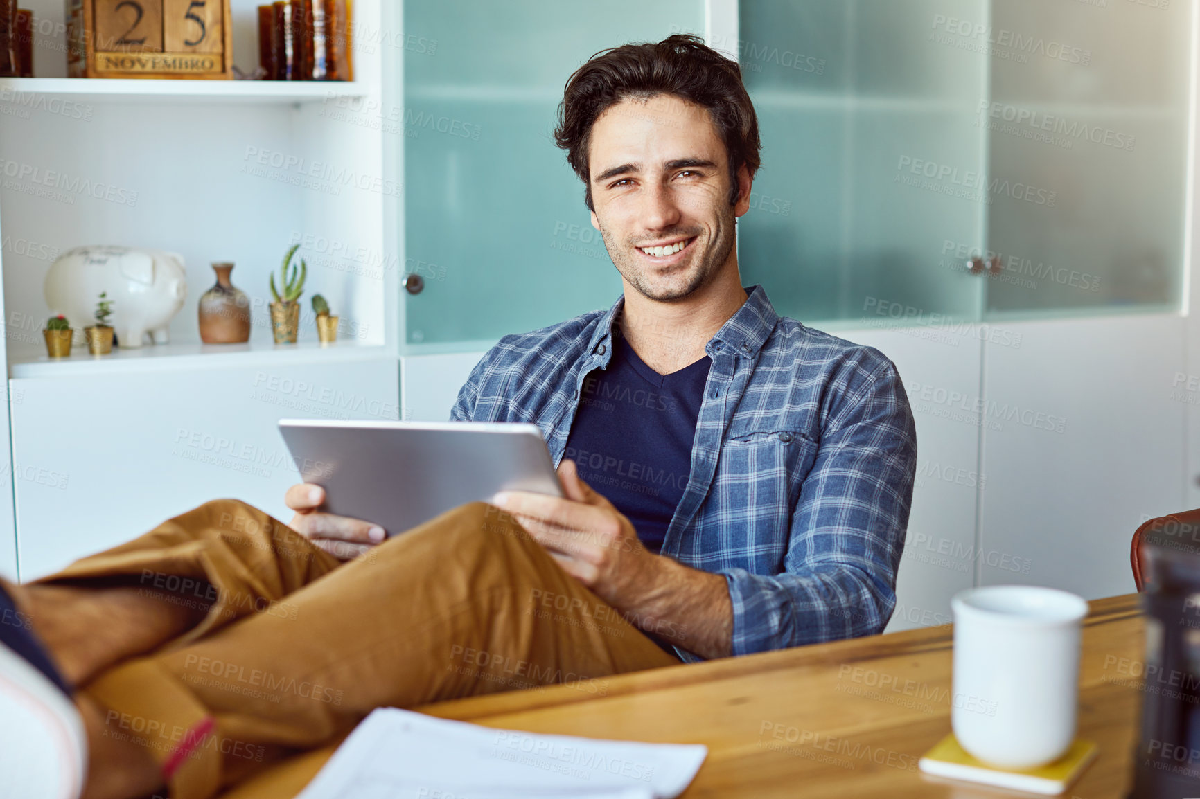 Buy stock photo Portrait of a cheerful young man working on a digital tablet while sitting with his feet up on a table at home