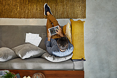 Buy stock photo High angle shot of a focused young unrecognizable man working on a digital tablet while relaxing on a couch at home